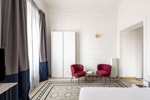 A hotel room with a bed, two red chairs, a small table, a TV, and a white door. - This beautiful apartment is located at Largo di Pietro Brazza 26, on the fourth floor. It is very bright and features two bedrooms, one of which has an en-suite bathroom. The spacious living room, an additional bathroom, and a fully equipped kitchen with (Roma)