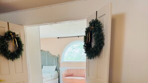 An open door leading to a bedroom with a bed, a pink chair, and a bench with flowers.