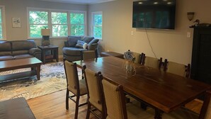 A living room with a wooden dining table, two sofas, a TV mounted on the wall, and a window with a view of trees.