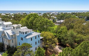 A large, multi-story building with a prominent blue facade, surrounded by lush greenery and overlooking a coastal area. - Chasing Sunsets in Seaside: 5 bikes, crow's nest (Santa Rosa Beach)