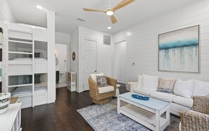 A spacious living room with a high ceiling fan, a white sofa set, a wicker chair, a coffee table, and a large painting on the wall. - Chasing Sunsets in Seaside: 5 bikes, crow's nest (Santa Rosa Beach)