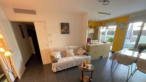 Una sala de estar con un sofá, una mesita con fruta, una zona de cocina con un fregadero y armarios, y un comedor con una mesa y sillas.