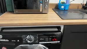 Uma cozinha com micro-ondas de aço inoxidável sobre bancada de madeira, um cesto azul com produtos de limpeza e uma máquina de lavar louça preta abaixo.