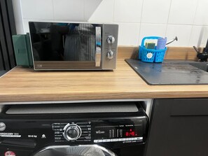 A kitchen with a stainless steel microwave on a wooden countertop, a blue basket with cleaning supplies, and a black dishwasher below. - 2-bed - Sleeps 5 - Free Parking - Town Centre (Grays)