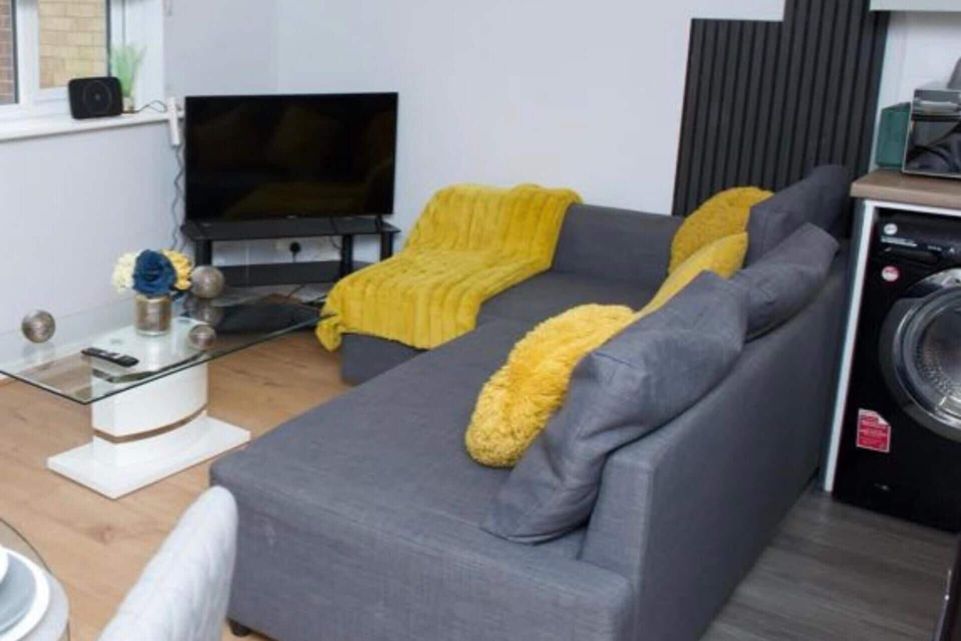 A living room with a grey sofa, a yellow throw blanket, a flat-screen TV, and a washing machine.