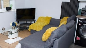 Sala de estar com um sofá cinza, um cobertor amarelo, uma TV de tela plana e uma máquina de lavar.