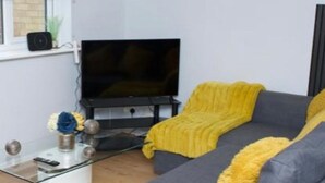 Una sala de estar con un sofá gris, un televisor de pantalla plana, una mesa de centro de vidrio y una manta amarilla.