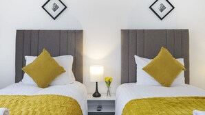 Habitación de hotel con dos camas, cada una con cabecero gris, ropa de cama blanca y una manta amarilla mostaza. Entre las camas, un mesita de noche con una lámpara y un jarrón con flores.