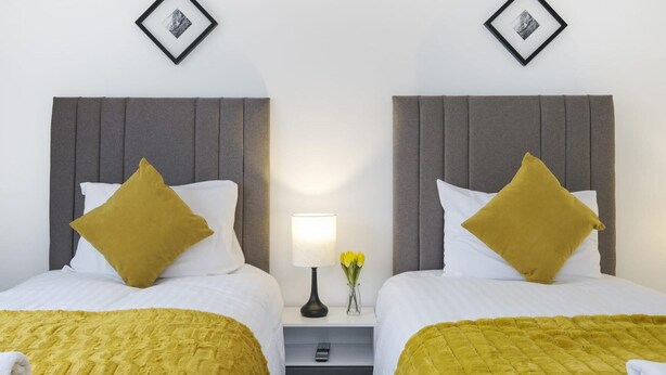 Quarto de hotel com duas camas, cada uma com cabeceira cinza, roupa de cama branca e manta amarela mostarda. Há uma mesinha de cabeceira com abajur e um vaso de flores entre as camas.