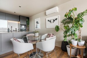 A modern kitchen with a dining area, featuring a round glass table, white chairs, and a large potted plant. - Stylish 1BD Flat & Car Park, Deck + Harbour Views (Auckland)