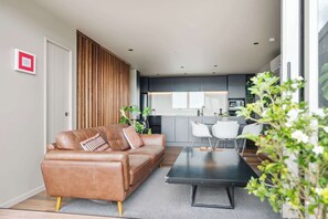 A modern living room with a leather sofa, a black coffee table, and a kitchen in the background. - Stylish 1BD Flat & Car Park, Deck + Harbour Views (Auckland)