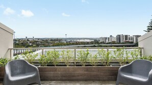 Een balkon met twee grijze stoelen, een houten plantenbak met groene planten en een stadsgezicht op de achtergrond.