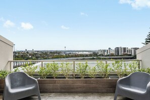 A balcony with two grey chairs, a wooden planter with green plants, and a cityscape view in the background. - Stylish 1BD Flat & Car Park, Deck + Harbour Views (Auckland)