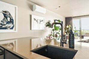 A modern kitchen with a stainless steel sink, a faucet, and a countertop. There are framed artworks on the wall and a potted plant in the background. - Stylish 1BD Flat & Car Park, Deck + Harbour Views (Auckland)