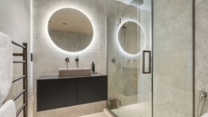 A modern bathroom with a freestanding sink, a glass shower enclosure, and a round mirror above the sink. - Stylish 1BD Flat & Car Park, Deck + Harbour Views (Auckland)