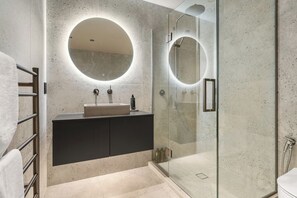A modern bathroom with a freestanding sink, a glass shower enclosure, and a round mirror above the sink. - Stylish 1BD Flat & Car Park, Deck + Harbour Views (Auckland)