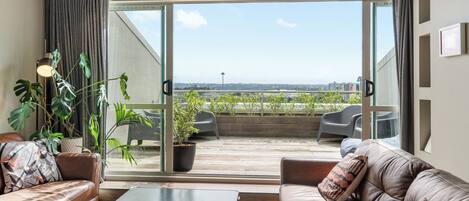Una sala moderna con un sofá, una mesa de centro y vistas a un balcón con plantas y un paisaje urbano.