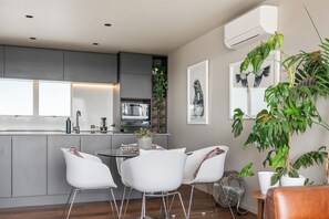 A modern kitchen with a dining area, featuring a central island, white chairs, and a large potted plant. - Stylish 1BD Flat & Car Park, Deck + Harbour Views (Auckland)