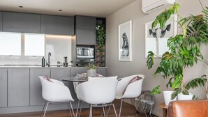 A modern kitchen with a dining area, featuring a central island, white chairs, and a large potted plant. - Stylish 1BD Flat & Car Park, Deck + Harbour Views (Auckland)