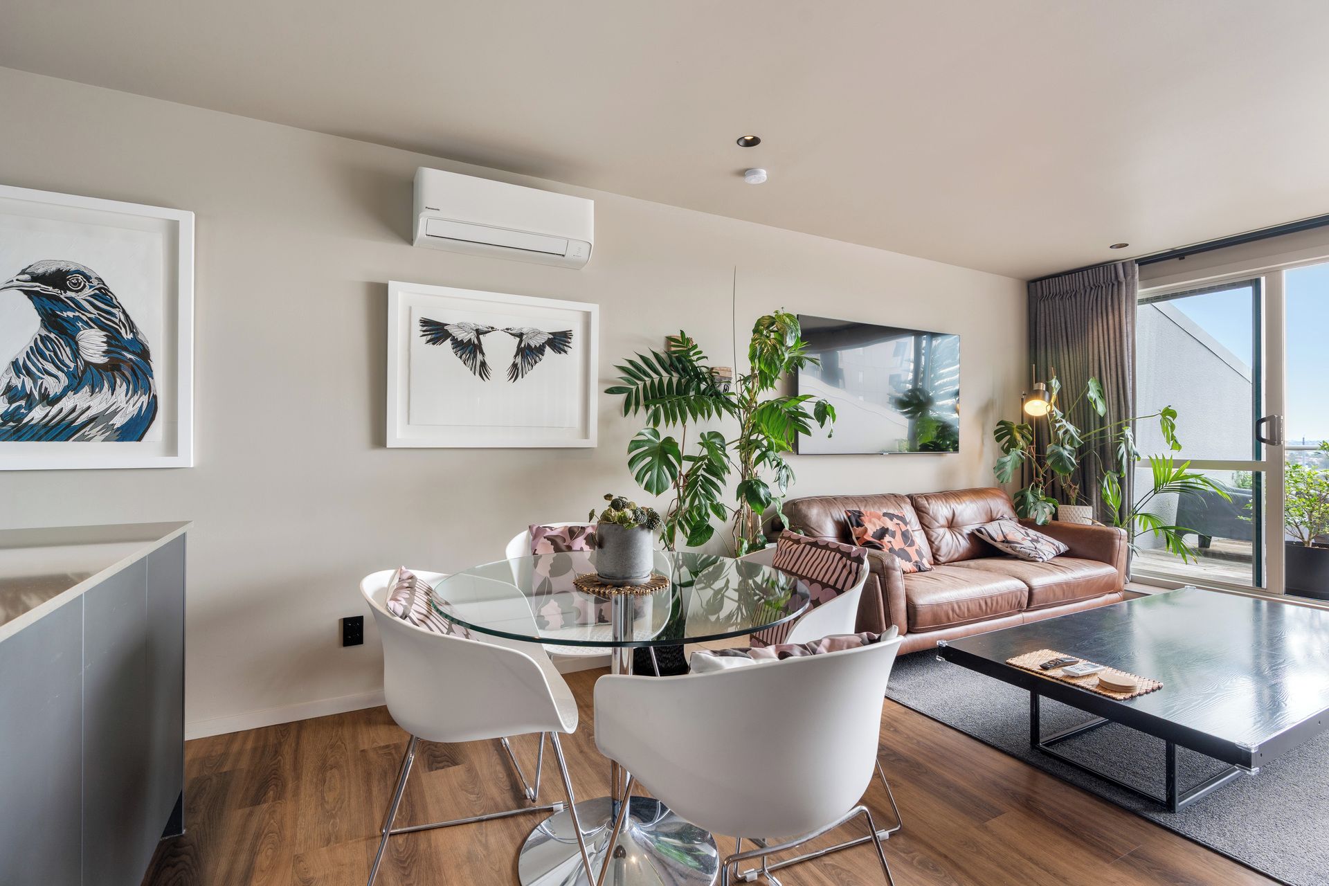 A modern living room with a glass dining table, white chairs, a leather sofa, and large windows.