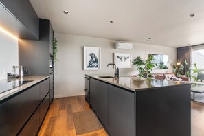 A modern kitchen with a central island, dark cabinetry, and a large window providing natural light. - Stylish 1BD Flat & Car Park, Deck + Harbour Views (Auckland)