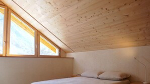 Ein Schlafzimmer mit schrägen Wänden, Holzbalken, einem Bett mit weißer Bettwäsche, einem Nachttisch und einem Fenster mit Blick zum Himmel.