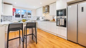 A modern kitchen with white cabinets, a stainless steel refrigerator, and a built-in oven. There are two black chairs with mesh backs, a wooden countertop, and a window with a view of houses. - Cozy Family & Contractor Home, 3X FREE Parking by Redwood Stays (Hampshire)