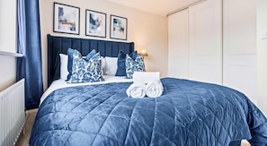 Un lit bien fait, recouvert d’une couverture bleu foncé, avec des oreillers blancs et une tête de lit bleue. Trois œuvres d’art encadrées sont accrochées au mur au-dessus du lit.