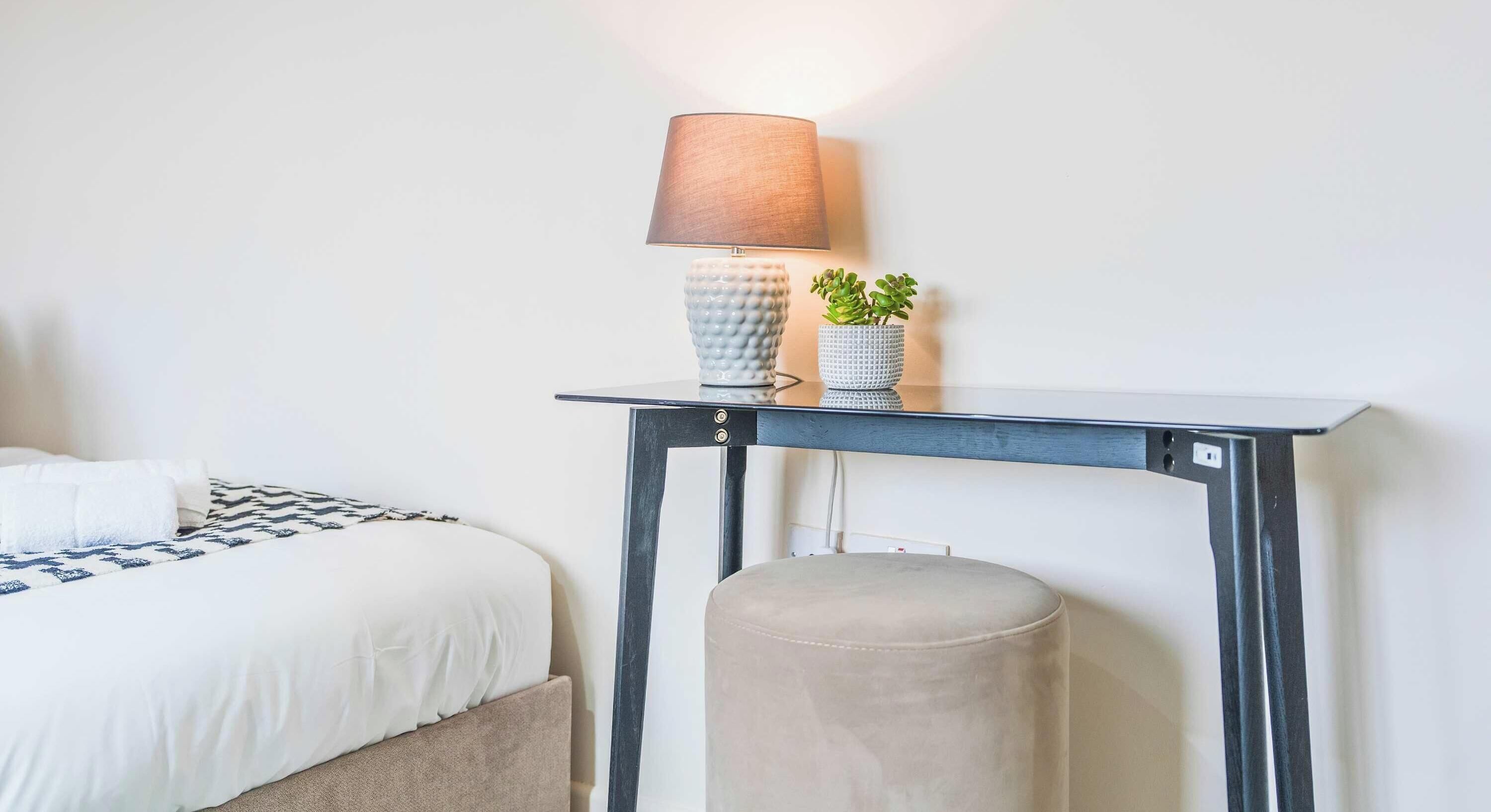 Un lit avec une couvre-lit à motifs, une table de chevet avec une lampe, une petite plante en pot et un pouf rond.