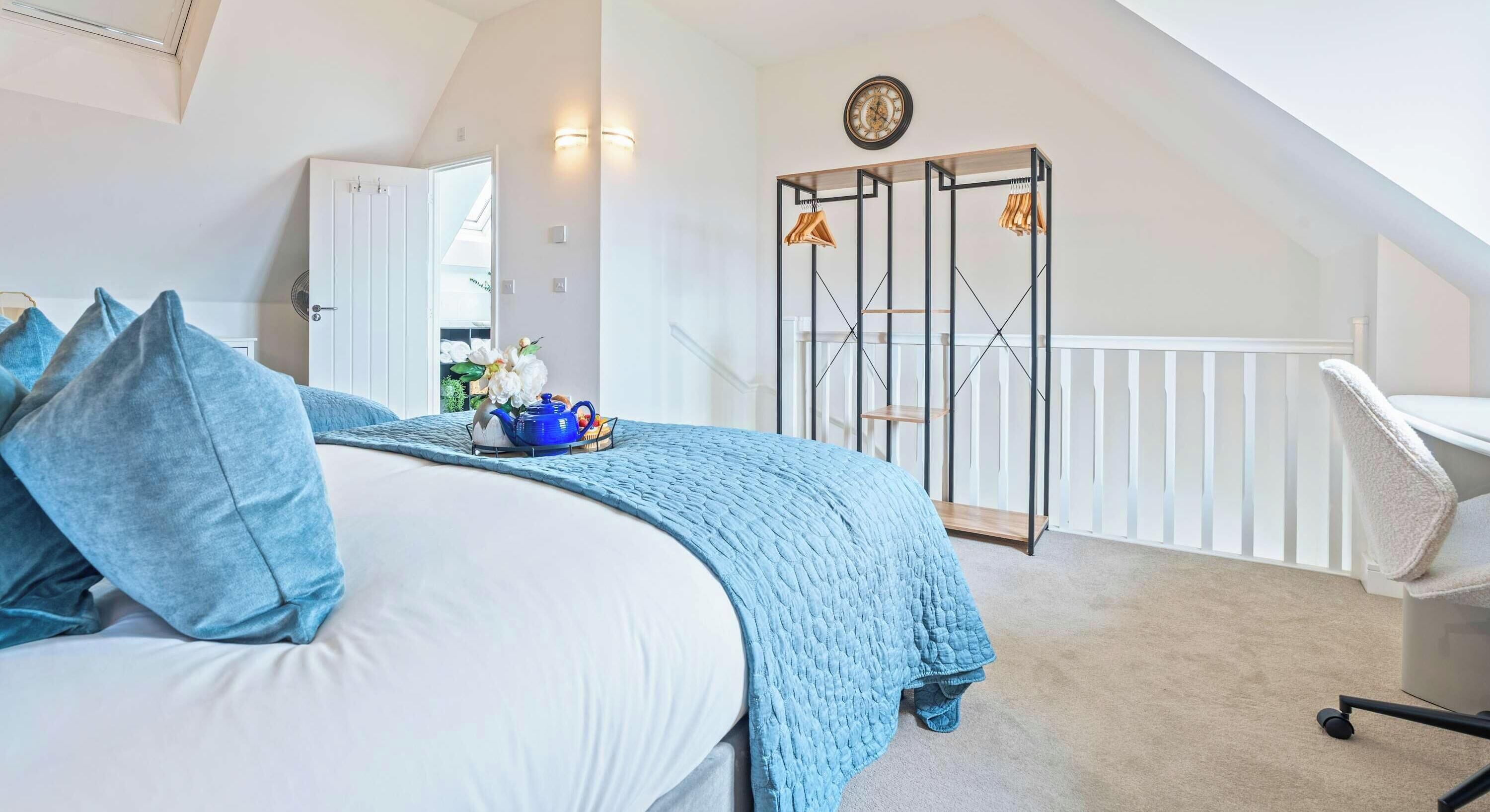 A bedroom with a bed, pillows, a blue blanket, a side table with a teapot, a chair, and a wardrobe with hanging clothes.