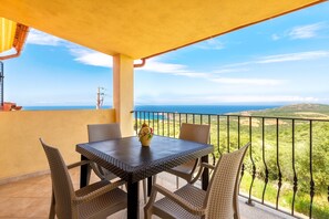 Un balcone con tavolo e sedie, un vaso con fiori e vista sul mare e le colline.