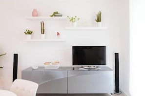 Ein modernes Wohnzimmer mit Flachbildfernseher, einer grauen Unterhaltungseinheit und weißen Regalen mit Pflanzen.