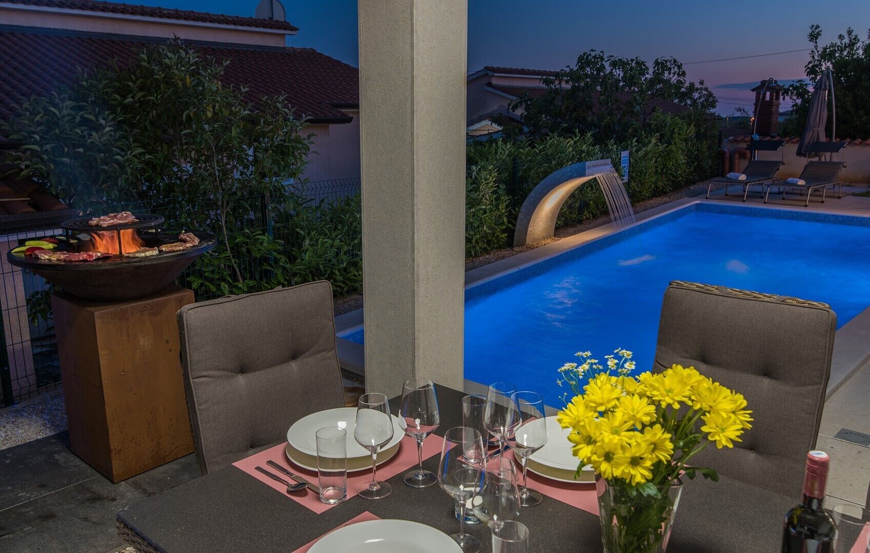 A poolside dining area with a table set for two, a grill, and a view of the pool.