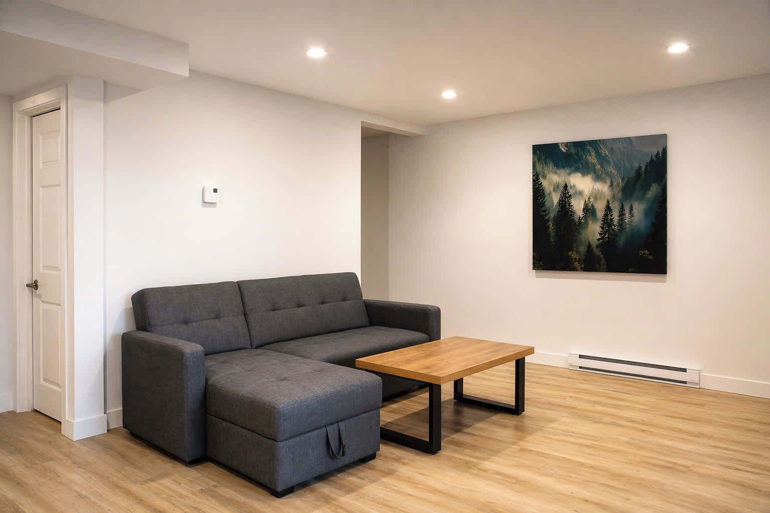 Ein modernes Wohnzimmer mit einem grauen Ecksofa, einem Holztisch und einem Waldgemälde an der Wand.