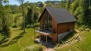 Una casa moderna de madera con balcón, rodeada de exuberante vegetación y un cielo despejado.
