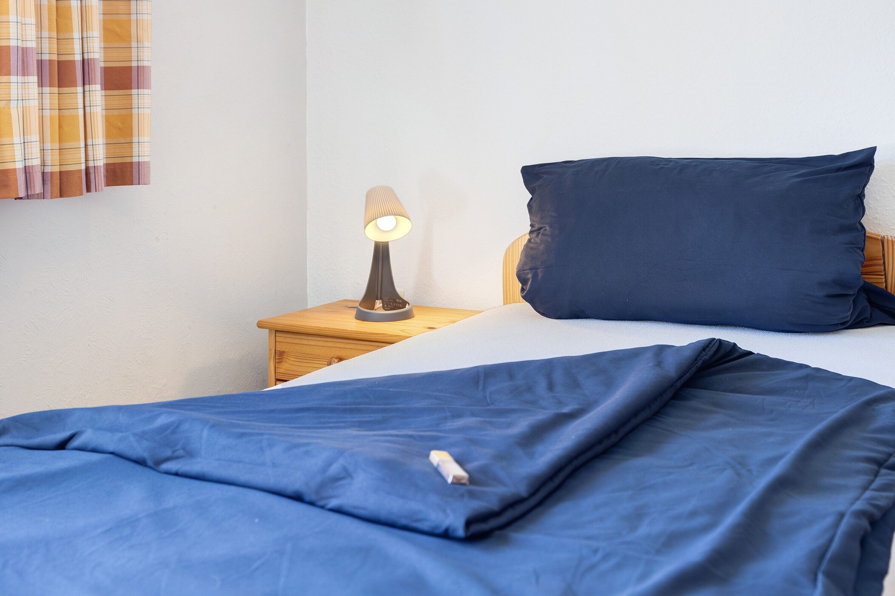 Ein Bett mit blauer Decke, einem dunkelblauen Kopfkissen und einer Nachttischlampe auf einem Holznachttisch.