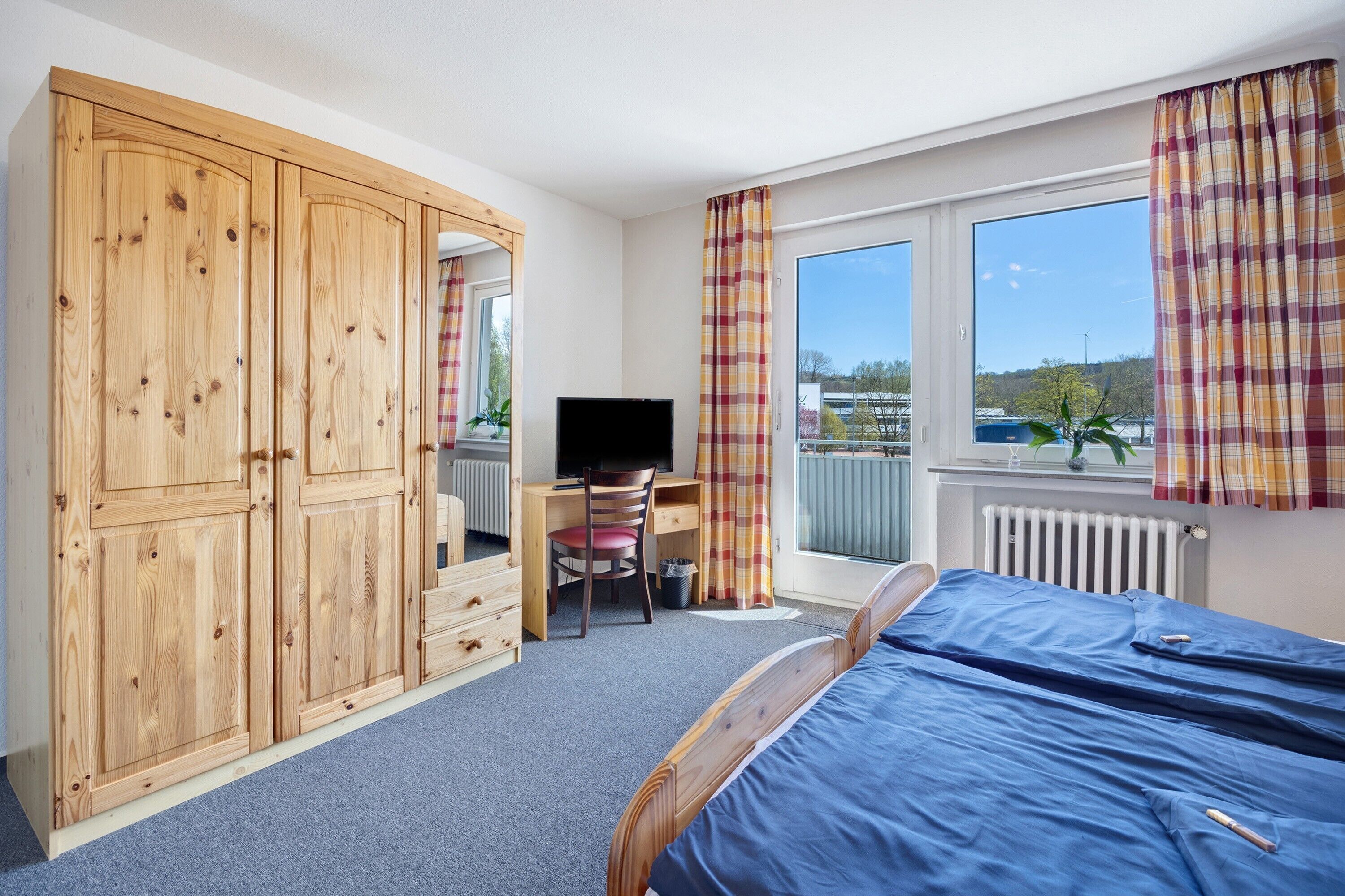 A bedroom with a bed, wooden wardrobe, desk, chair, television, and large windows with curtains.