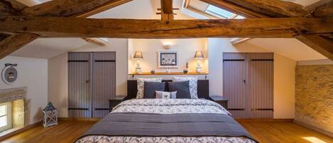 A bedroom with a sloped ceiling, wooden beams, a bed with floral bedding, bedside lamps, a small table, and a wall-mounted shelf with decorative items.