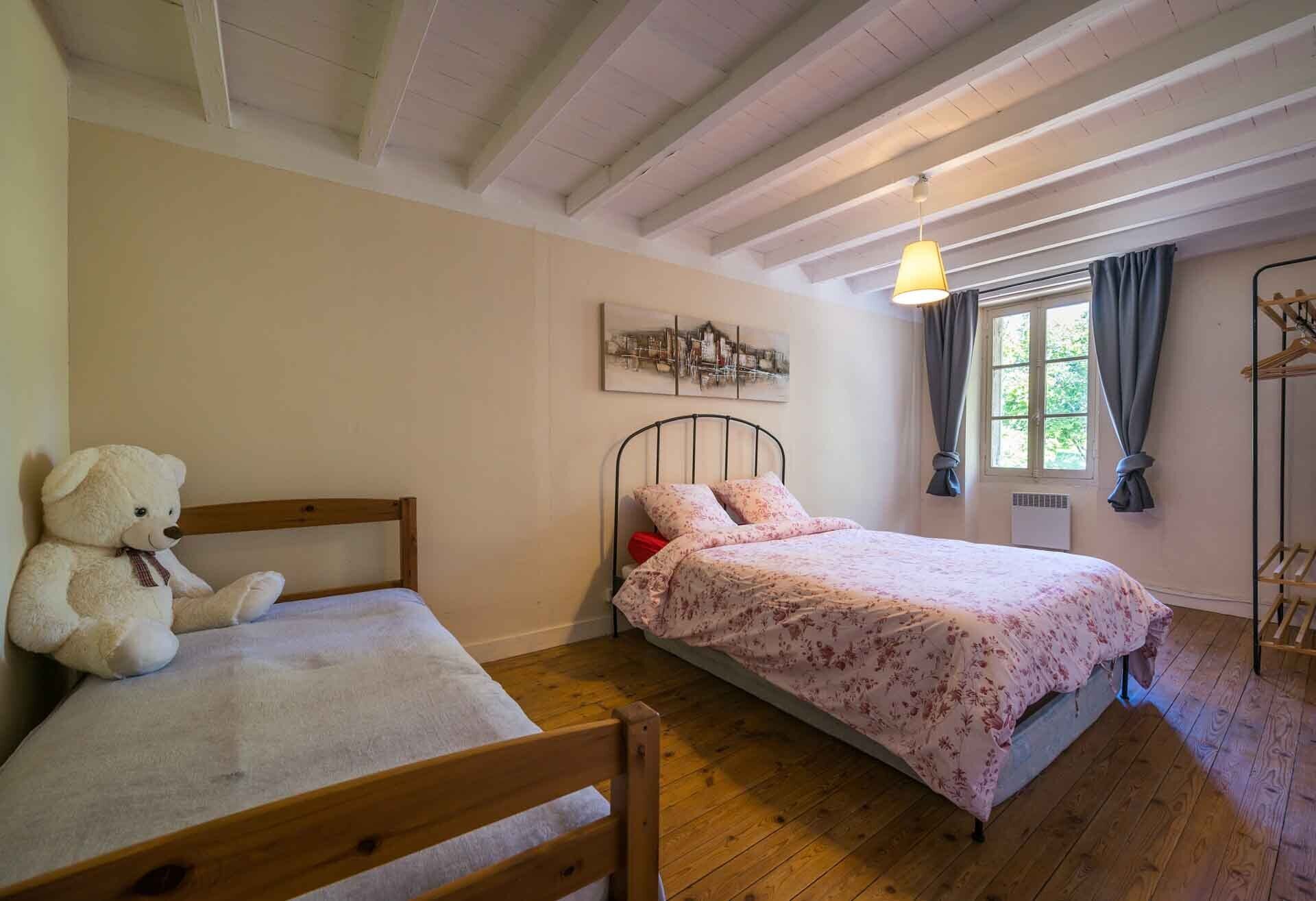 Une chambre à coucher avec un lit, une chaise, un ours en peluche, une fenêtre et un luminaire suspendu.