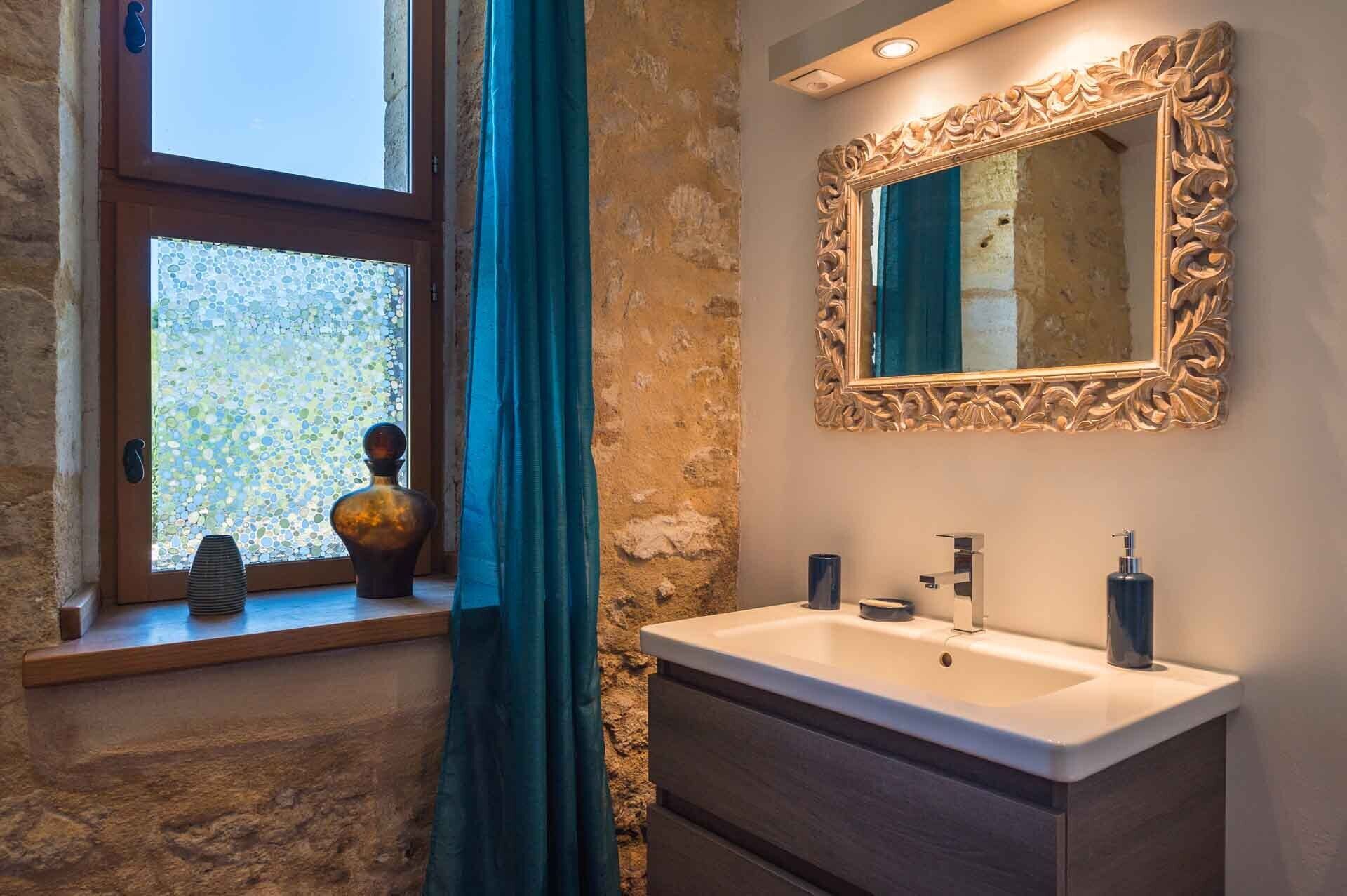 Une salle de bain avec un mur en pierre, une fenêtre offrant une belle vue, un miroir doté d’un cadre orné et un lavabo avec des accessoires modernes.
