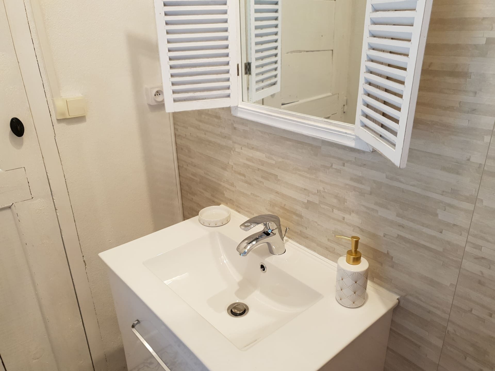 Une salle de bain avec un lavabo blanc, un miroir avec des volets blancs ouverts et un mur recouvert de carreaux beiges.