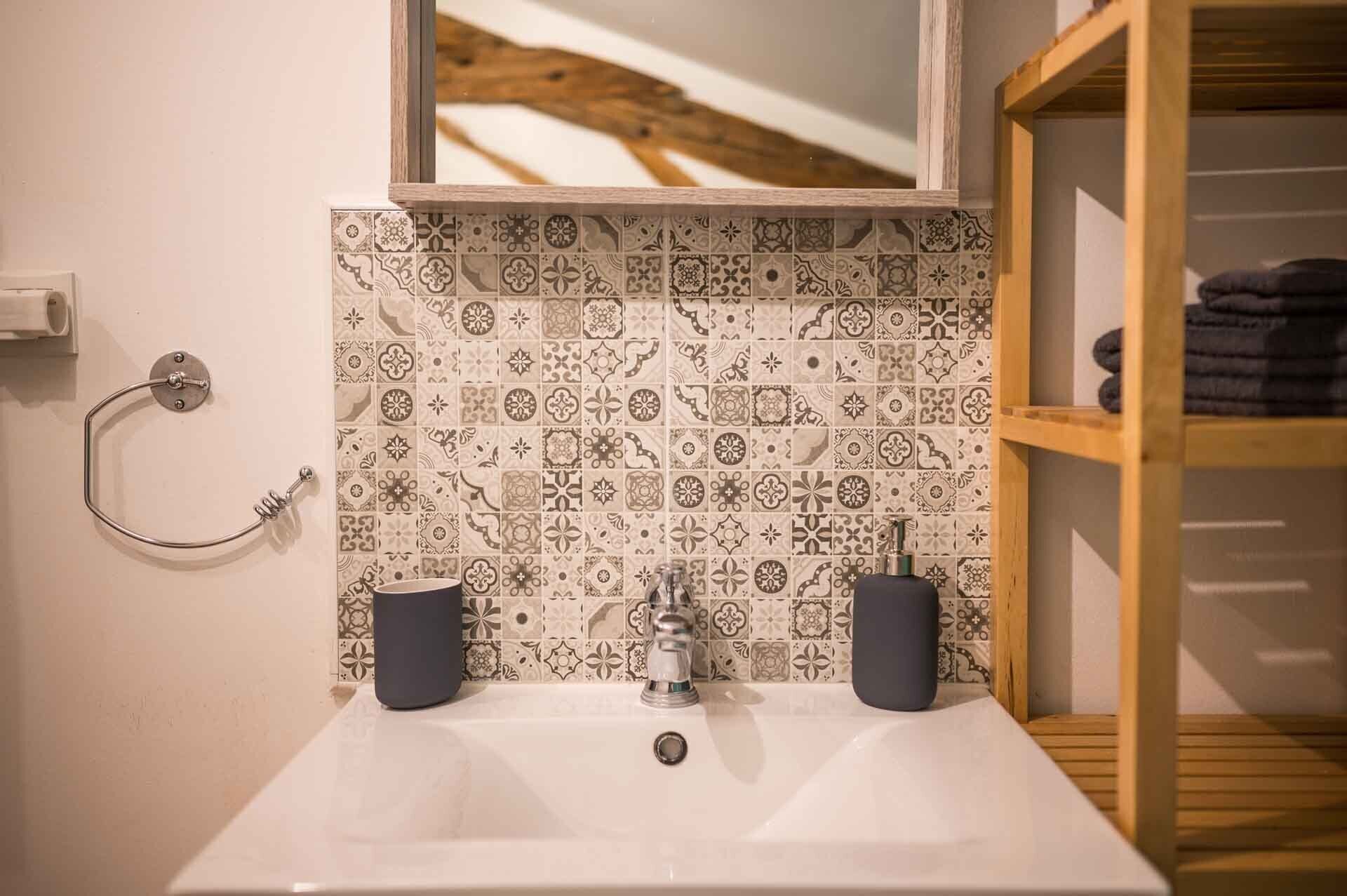 Une salle de bain avec un lavabo blanc, des carreaux à motifs, un miroir et une étagère en bois.