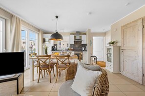 A modern kitchen with white cabinets, a dining area with wicker furniture, and a TV on a wooden stand. - The Beach House: beach villa with sauna, whirlpool and roof terrace in Olpenitz (Kappeln)