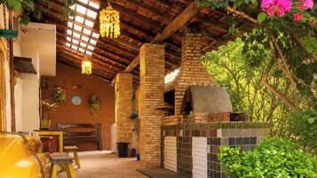 An outdoor patio area with a brick oven, wooden beams, hanging lights, and potted plants.