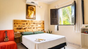 A bedroom with a wooden headboard, a bed with a white bedspread, a red chair, a guitar, and a window with shutters. - Casa Vilar Rio Lençois Maranhenses (Barreirinhas)