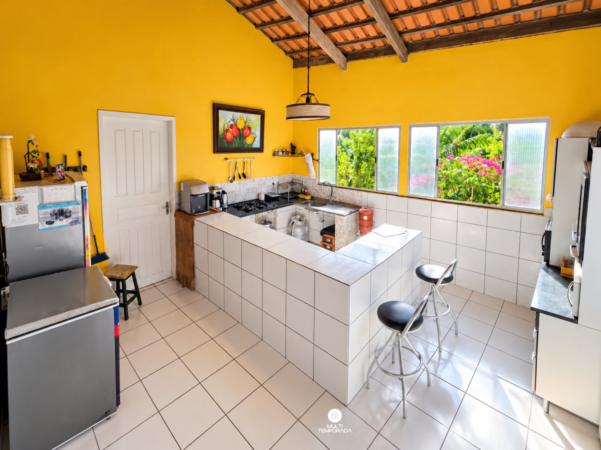 A compact kitchen with a central island, white tiles, and a view of greenery through large windows.