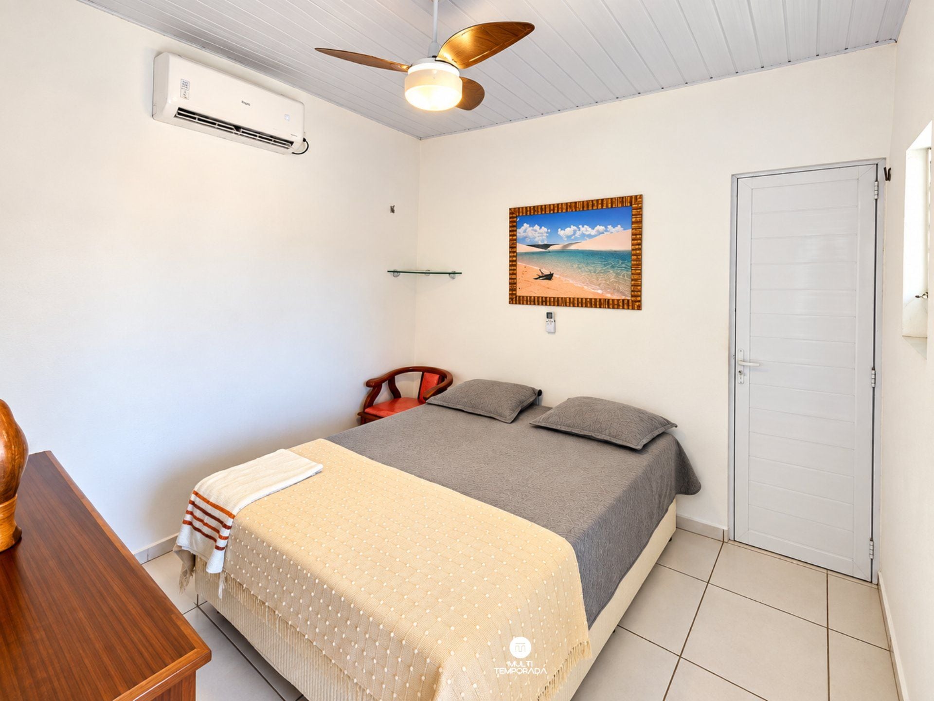 A bedroom with a bed, a chair, an air conditioning unit, a ceiling fan, a door, and a wall-mounted picture.