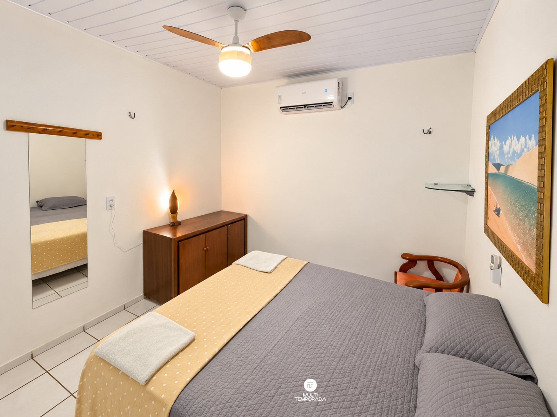 A bedroom with a bed, a wooden dresser, a mirror, an air conditioner, a ceiling fan, and a painting on the wall.