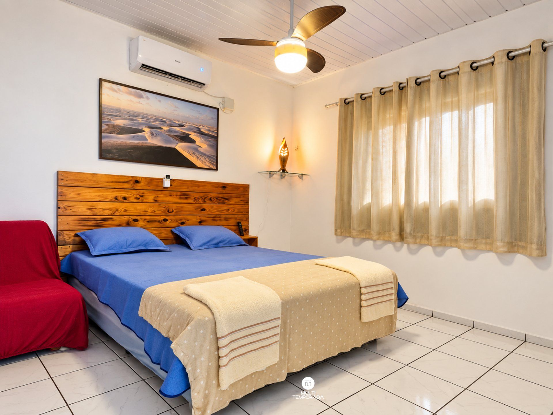 A bedroom with a wooden headboard, a bed with blue bedding, a red chair, a ceiling fan, an air conditioning unit, a wall-mounted lamp, and a window with curtains.