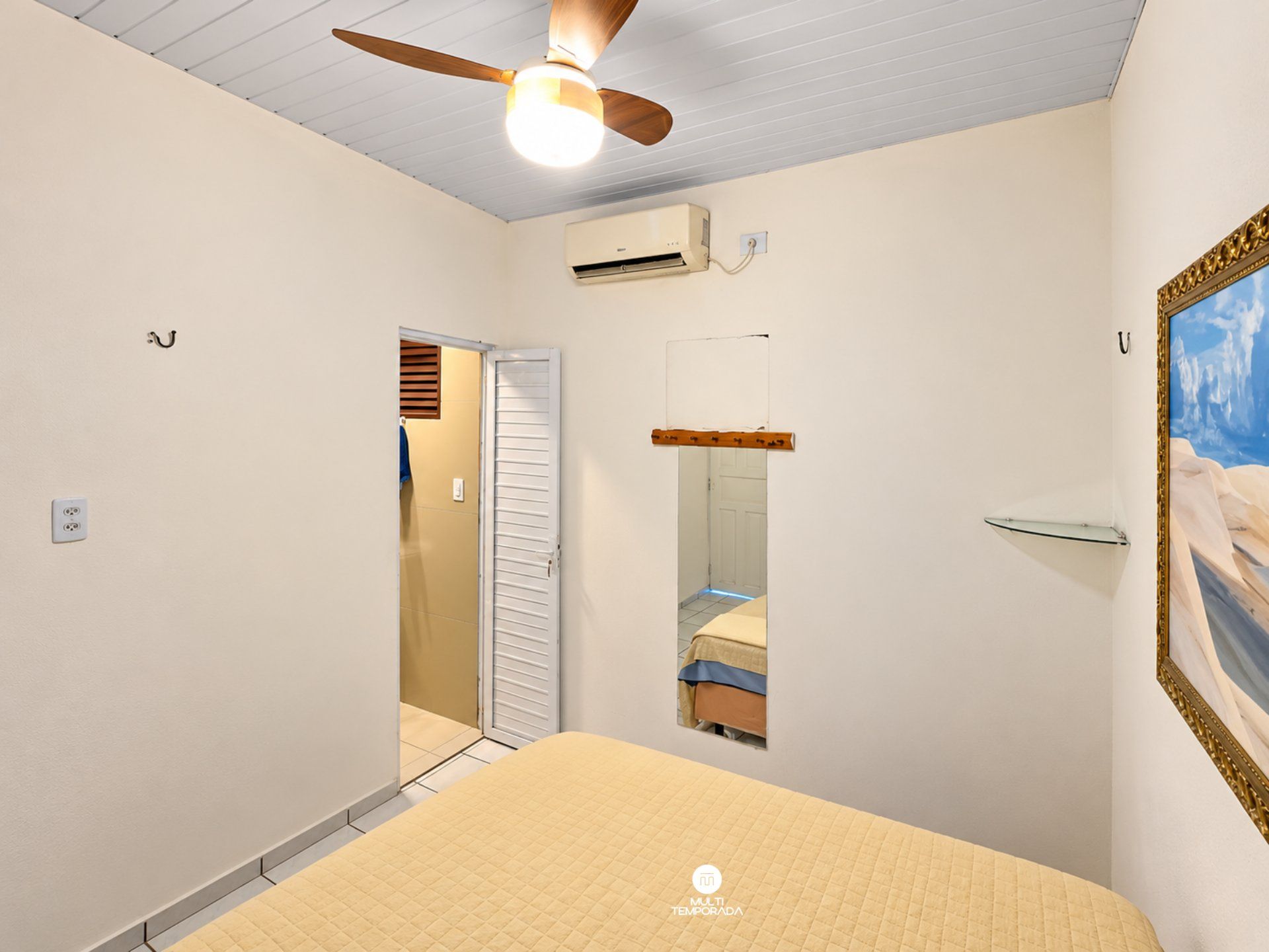 A bedroom with a bed, a ceiling fan, an air conditioning unit, a door, and a painting on the wall.